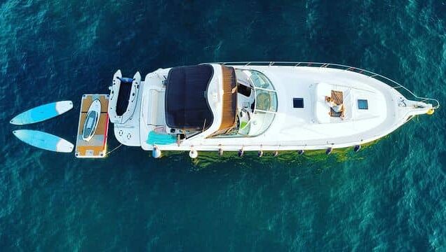 Yate blanco anclado en aguas cristalinas, con dos tablas de paddle surf en el lado y una persona relajándose en la cubierta.