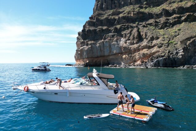 Grupo de personas disfrutando de un día en el mar, nadando y relajándose en una lancha y una plataforma flotante, con acantilados de fondo y un cielo despejado.